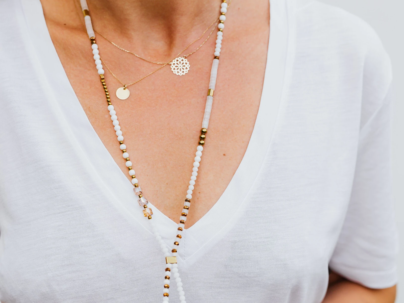 Woman in a white V-neck tee wearing a long white and gold beaded necklace layered with a fine gold pendant necklace