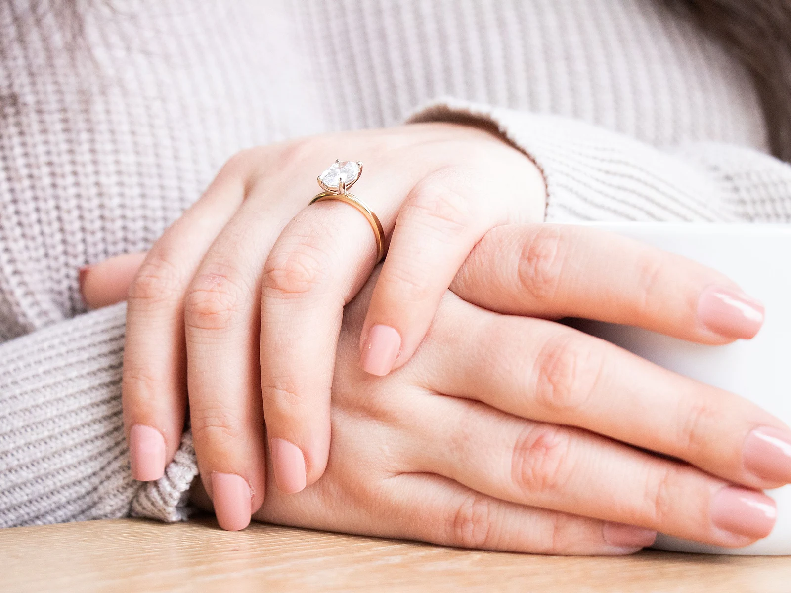Hands in a cozy knit sweater wearing a solitaire diamond ring beside a coffee mug