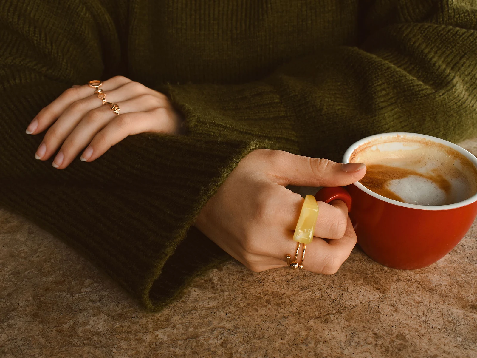 Hands in an olive knit sweater wearing stacked gold rings while holding a latte