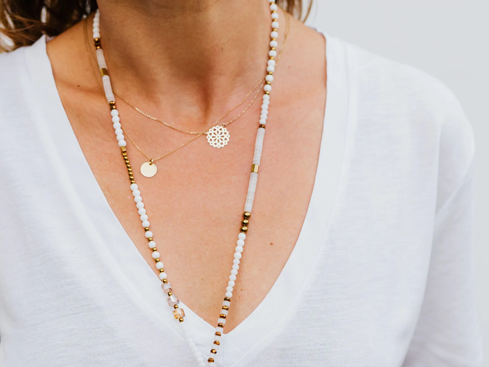 Woman in a white V-neck T-shirt wearing two fine gold pendant necklaces layered with a long beaded necklace