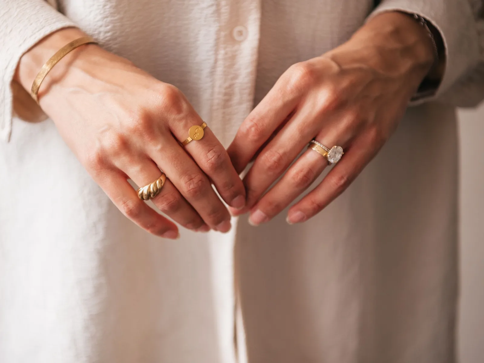 Two hands resting together wearing thin gold rings and a diamond solitaire