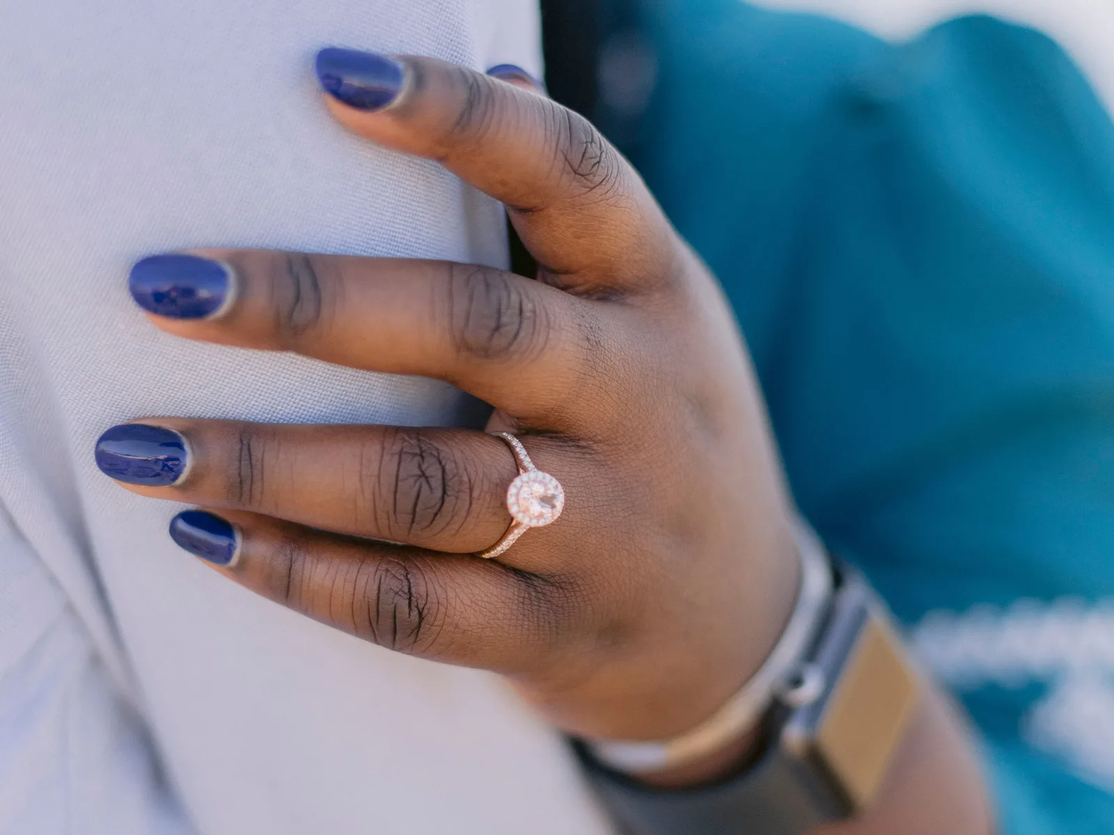 Rose gold halo engagement ring with an oval center stone worn on a hand with dark blue nail polish